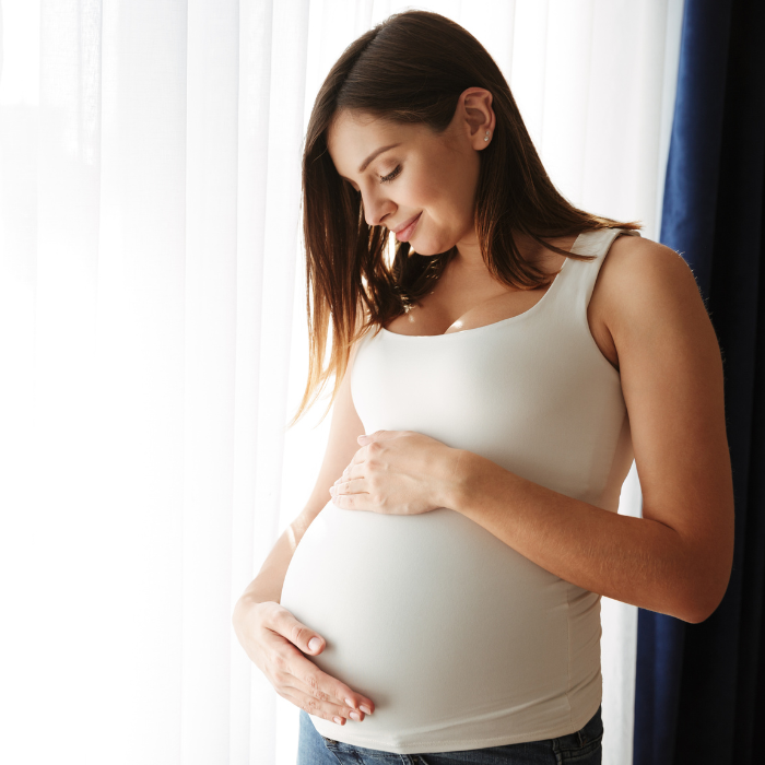 Pregnant woman in white tank top stands by curtain smiling at her belly.